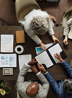 Group of middle aged multiethnic business professionals collaborating around table, reviewing documents and using laptop, top view showing teamwork and corporate meeting environment