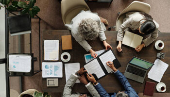 Group of middle aged multiethnic business professionals collaborating around table, reviewing documents and using laptop, top view showing teamwork and corporate meeting environment