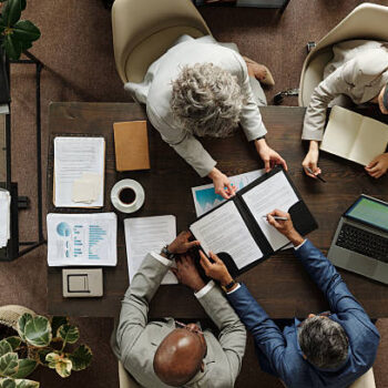 Group of middle aged multiethnic business professionals collaborating around table, reviewing documents and using laptop, top view showing teamwork and corporate meeting environment